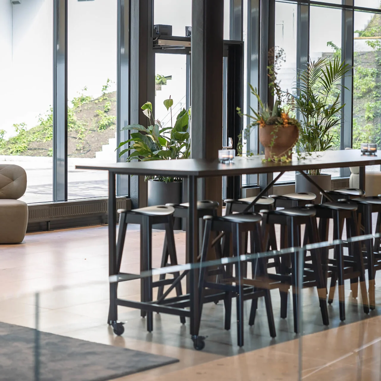 Barstole omgiver et højt bord i en moderne, lys lounge med store vinduer og grønne områder udenfor. Komfortable stole og potteplanter forbedrer den indbydende atmosfære.