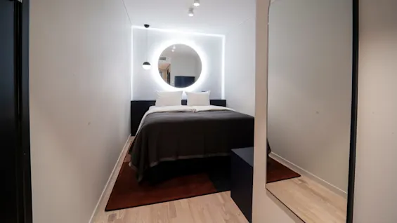 A bed with white pillows and dark blanket, placed against a wall with a circular backlit mirror. The room is minimalistic, with light wood floors and a hanging lamp.