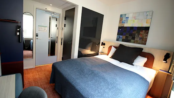 Moderni hotellihuone, jossa on sijattu parivuode, värikästä taidetta seinällä ja valoisa käytävä taustalla.
