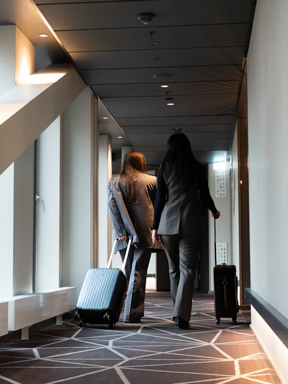 Två kvinnor i kostym går längs en hotellkorridor med geometrisk matta och drar rullväskor efter sig.
