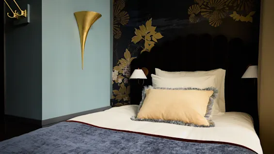 A neatly made bed with decorative pillows sits against a dark floral-patterned wall, featuring two wall lamps and a golden cone-shaped decoration. The surrounding walls are pale green.