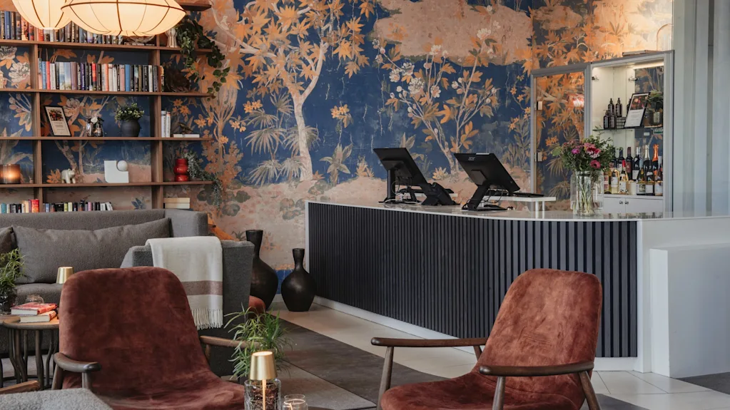En hotellounge med servicedisk, bar og et hyggeligt siddeområde. Et levende vægmaleri af træer og løvværk pryder væggen.