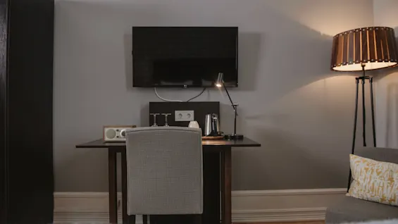A desk with a chair, lamp, kettle, and radio is positioned against a gray wall with a mounted TV. A tall lamp stands to the side; cozy room setting.