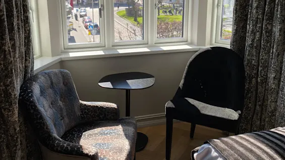 Two armchairs face each other with a small round table between them in a sunlit room. Large windows reveal a scenic view of buildings, trees, and a hill outside.