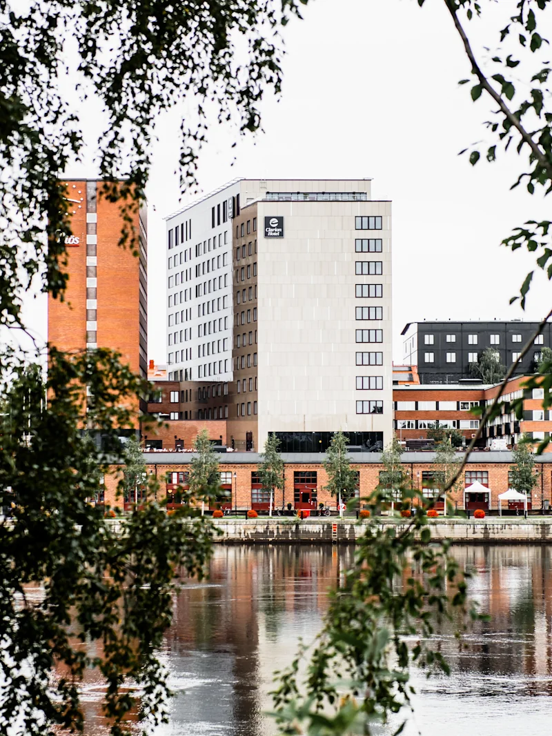 Høye bygninger står tvers over en elv, sett gjennom løvrike tregrener, og skaper et innrammet bylandskap.