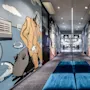 Hallway adorned with large, colorful murals of cartoon figures in suits, leading to a well-lit, open area. Blue cushioned benches sit on grey carpet flooring.