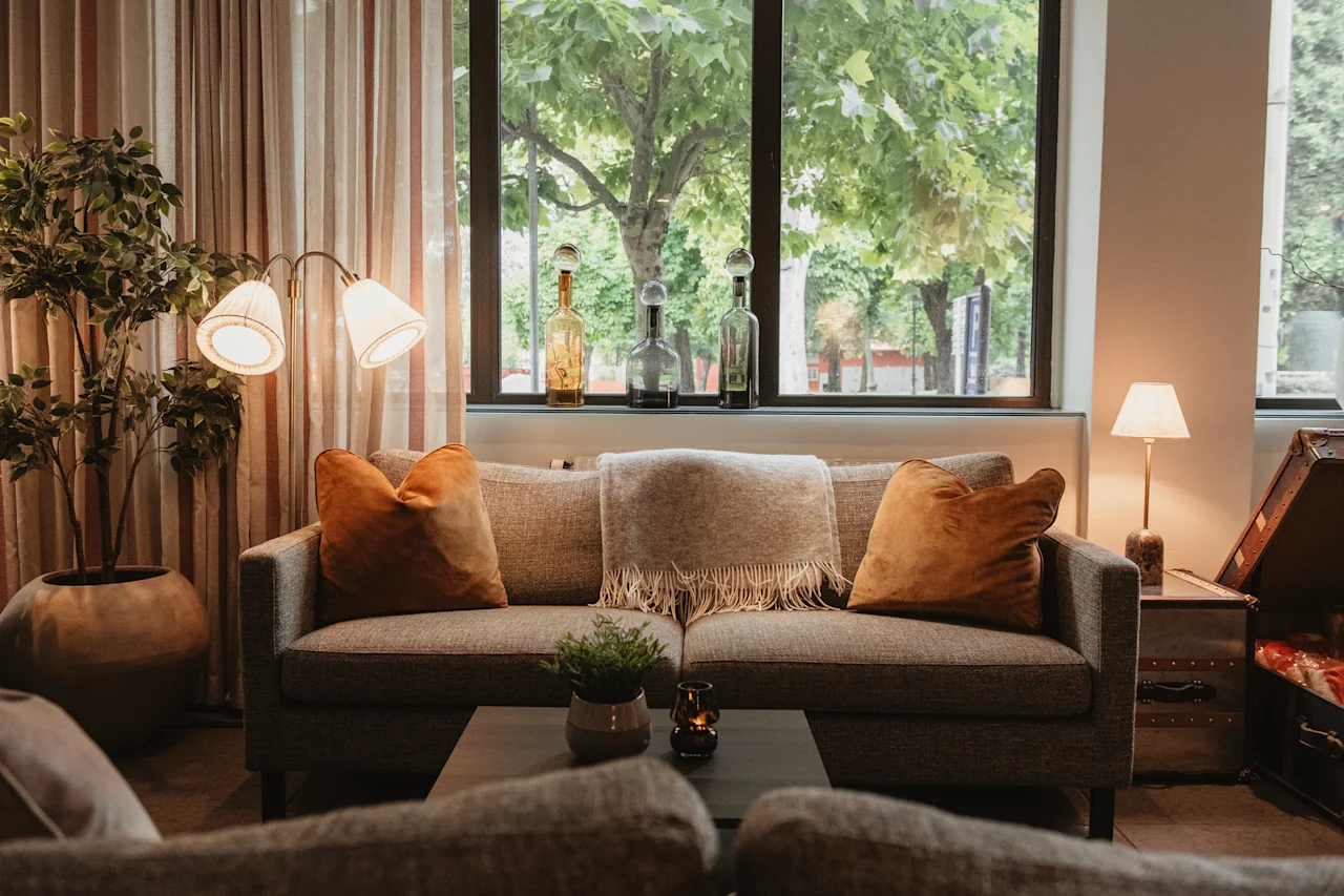 A cozy lounge area with a sofa, warm lighting, and lush greenery visible through a large window, creating an inviting atmosphere.