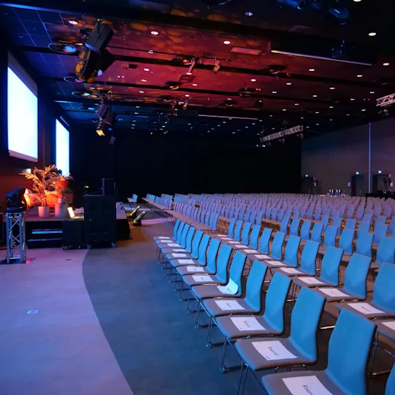 Rader av tomma blå stolar med "Reserved"-skyltar, i en rymlig konferenshall med stora skärmar, scen och omgivande belysning. Växtdekorationer på sidan.
