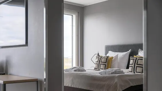 A neatly made bed with geometric-patterned pillows and folded robes is positioned under a large window, providing natural light in a modern, gray-walled bedroom.