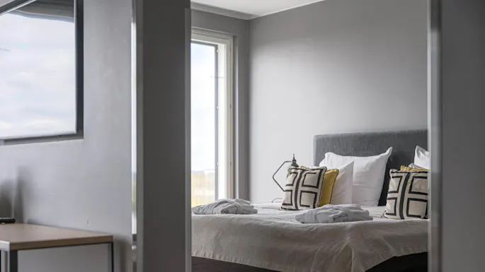 En pent og ryddig seng med puter i geometriske mønstre og sammenbrettede badekåper er plassert under et stort vindu som gir naturlig lys i et moderne soverom med grå vegger.