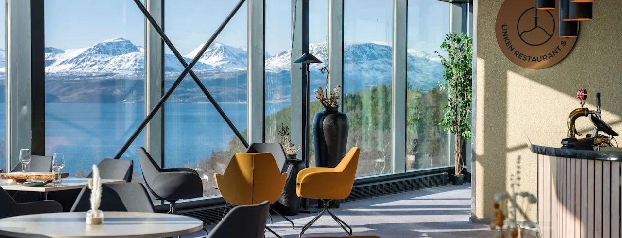 Bord och stolar står i en modern restaurang med panoramafönster som erbjuder utsikt över snötäckta berg och vatten. En skylt lyder, "LINKEN RESTAURANT &" på väggen.
