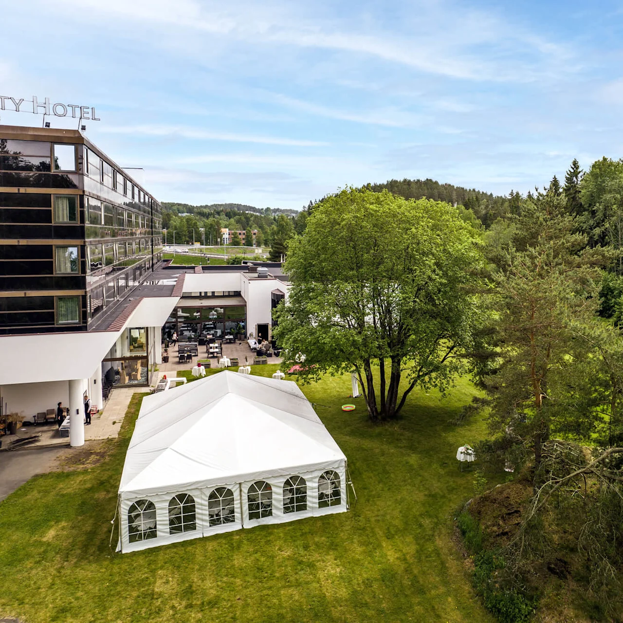 A large white event tent stands on green grass beside a modern hotel building, surrounded by lush trees under a clear sky. Partial text on the building reads: "ITY HOTEL."