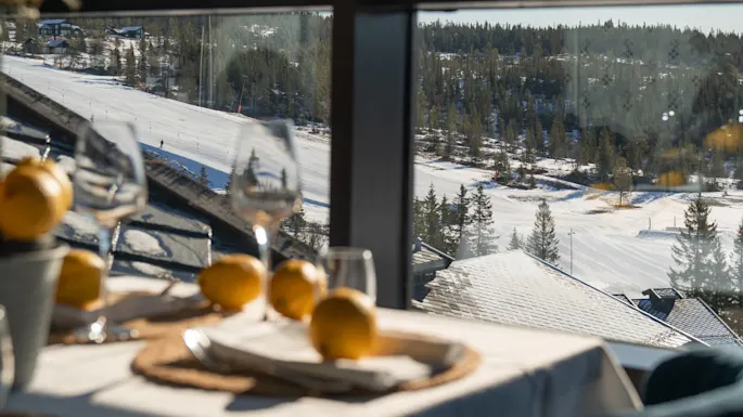 Table set with lemons and wine glasses, beside a large window. Outside, a snowy ski slope stretches across a forested landscape under a clear blue sky.