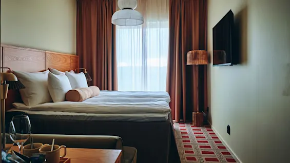 Et koselig hotellrom med en komfortabel seng, et stort vindu med gardiner, en veggmontert TV og et nattbord.