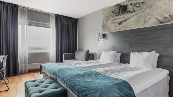 A modern hotel room with two beds, a teal throw, and a large window with dark curtains, offering a comfortable stay.