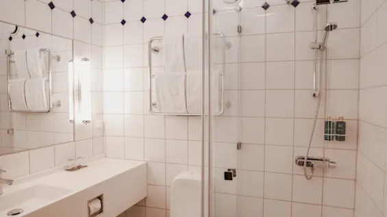 Bathroom featuring a white sink and mirror; white towels hang on a rack. Showerhead, shampoo, and conditioner are set against tiled walls with dark diamond accents.