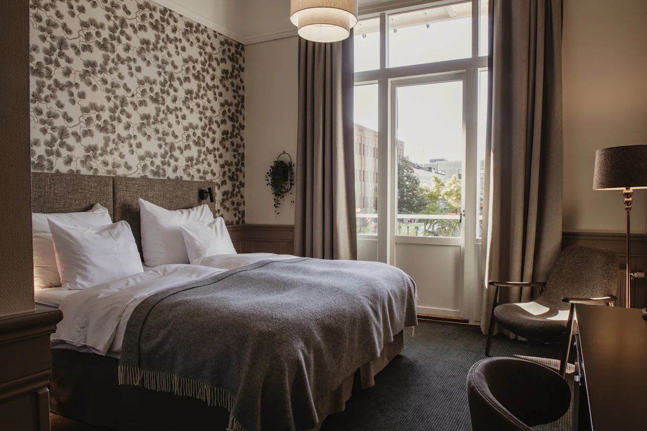 Ett bekvämt hotellrum med en stor säng med vita lakan, blommig tapet och ett ljust fönster med gardiner.