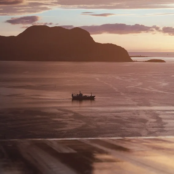 En båt som seiler over blikkstille vann i solnedgang med en mørk fjellsilhuett i bakgrunnen.