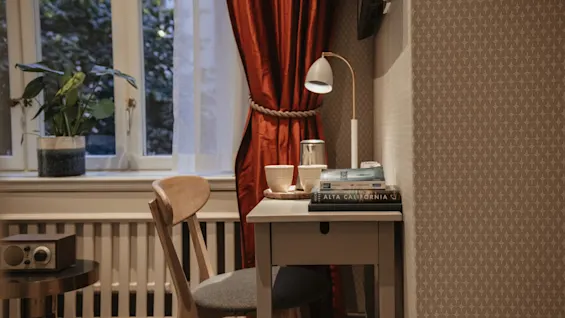 Et hyggeligt skrivebordsområde på Home Hotel Wellington med en lampe, bøger inklusive ALTA CALIFORNIA, og et kaffeopsætning ved siden af et vindue med røde gardiner.