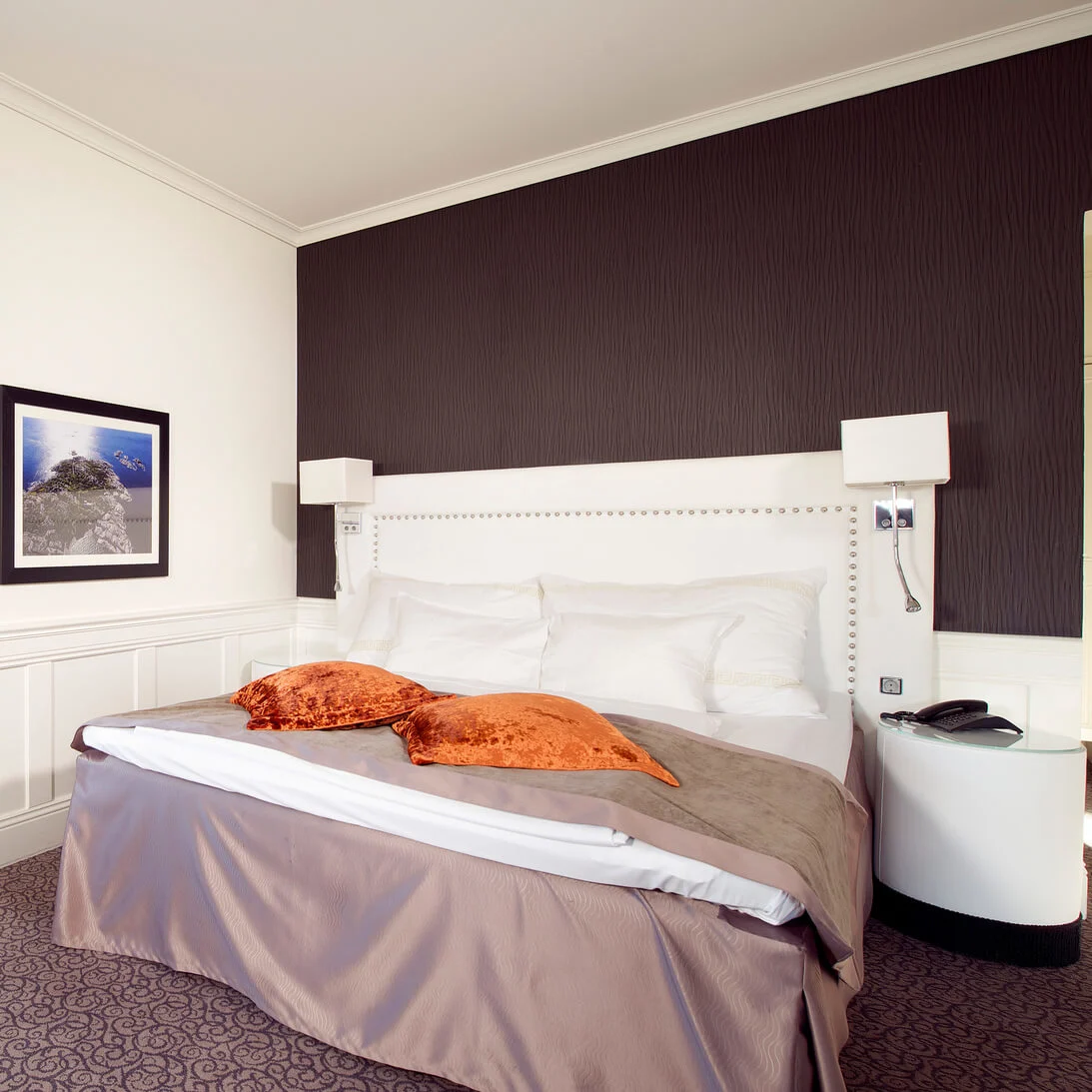 En dubbelsäng med vita kuddar och orange kuddar står mot en mörk fondvägg, flankerad av sängbord, i ett hotellrum med ett fönster och inramad väggkonst.
