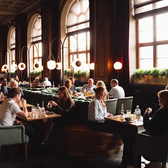 Personer äter och samtalar i en rymlig, elegant designad restaurang med höga fönster, moderna cirkulära lampor och frodig grönska på fönsterbrädorna. Atmosfären är livlig och välkomnande.