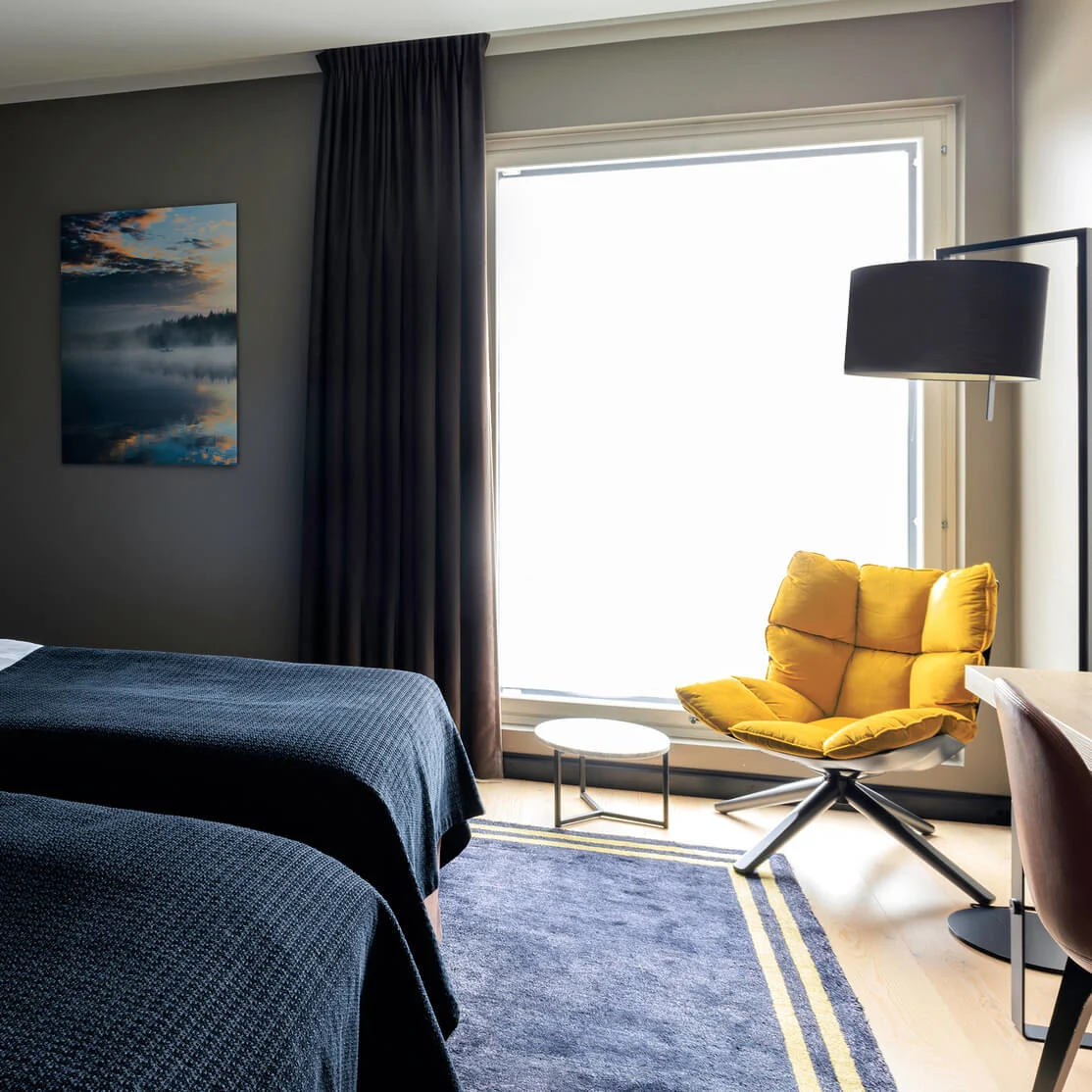 Two neatly made beds with green pillows face a large window. A cozy yellow chair and small table sit beside, with a painting of a misty lake on the wall.
