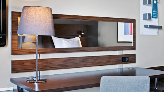 En moderne skrivebordslampe lyser op i et spejl i hotelværelset og reflekterer en pude. På væggen hænger et skilt med "Venligst ikke forstyrre".
