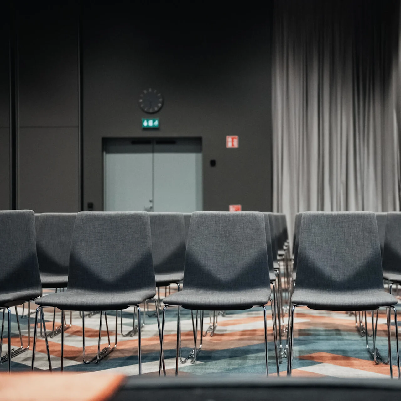 Rader av tomma grå stolar vända mot en stängd dörr med utgångsskyltar och en klocka, i ett svagt upplyst konferensrum.