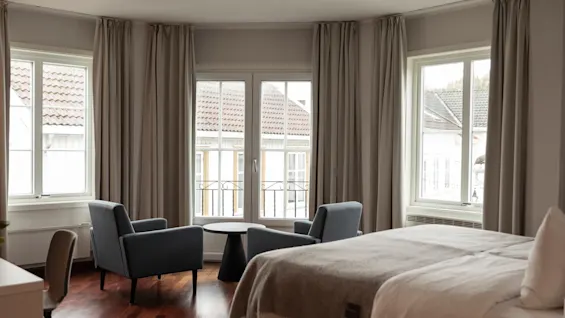 A cozy hotel room with a bed, two armchairs, and a small table by the windows, offering a view of buildings outside.