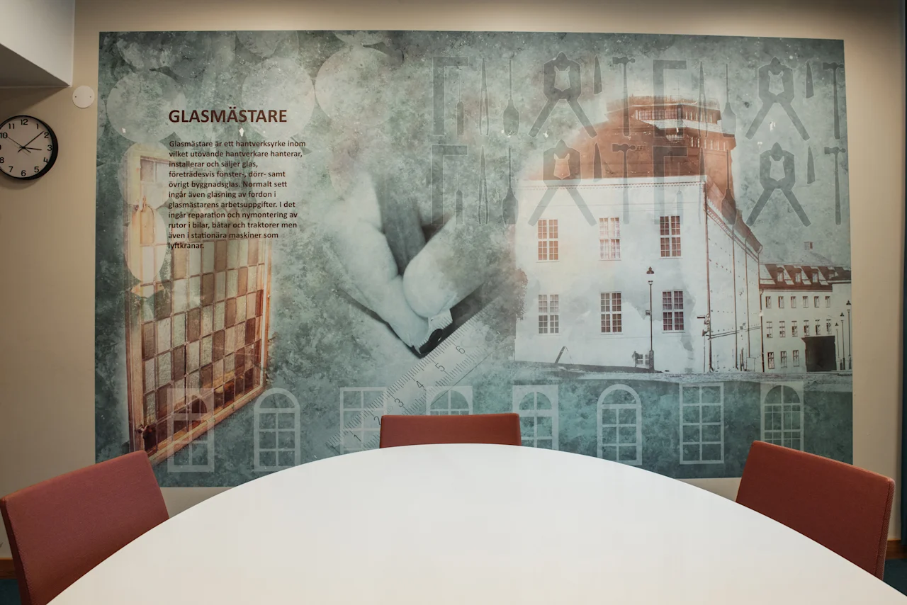 Mural of glazier craft with text GLASMASTARE, hands, tools, and buildings, above a round table. Home Hotel Uppsala.
