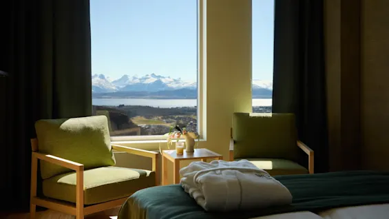 Two green armchairs face large windows revealing snow-capped mountains and a lake. A wooden table with decorative items sits between the chairs. A folded robe lies on a bed.