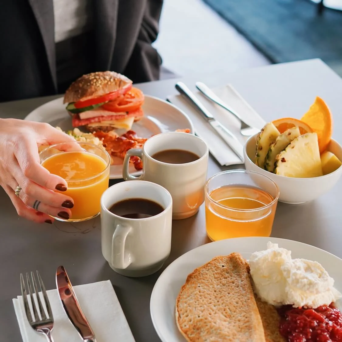 A person holds a glass of orange juice over a breakfast table with coffee, fruit, toast, and a sandwich. The setting is bright and modern.