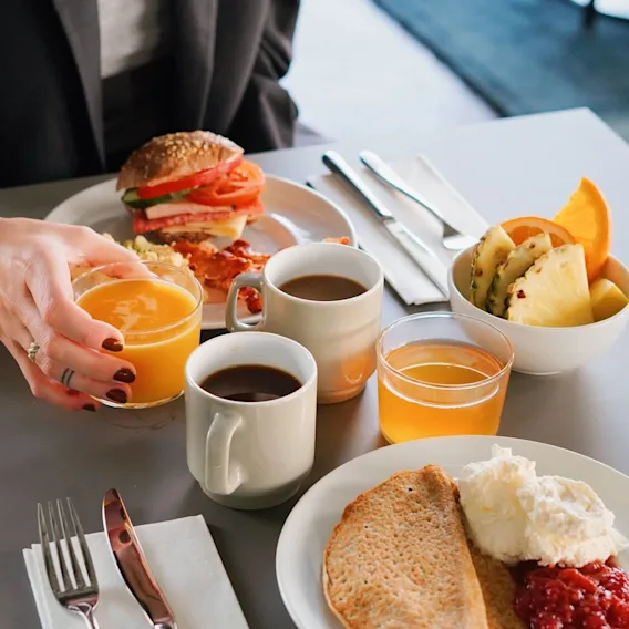 En person håller ett glas apelsinjuice över ett frukostbord med kaffe, frukt, toast och en smörgås. Miljön är ljus och modern.