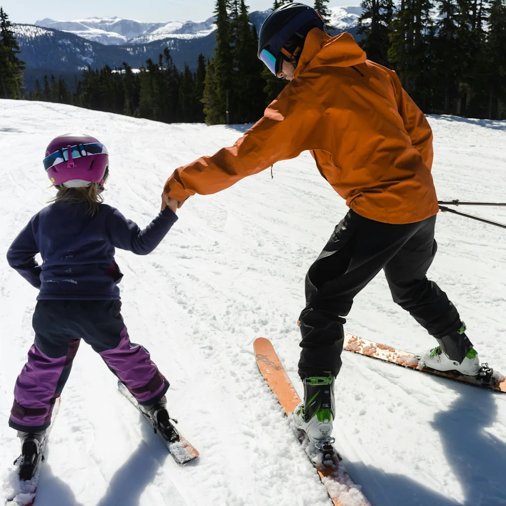 An adult skier holds a child's hand on a snowy mountain slope, teaching them to ski with trees and mountains in the background.