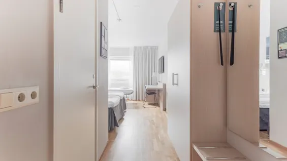 A bright hotel room interior with a hallway leading to two beds, a desk, and a window. A wardrobe with do not disturb signs.