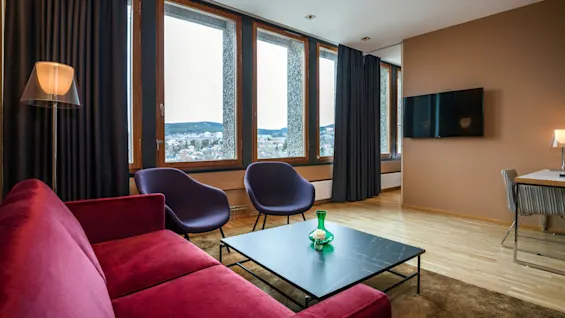 Red sofa and two purple chairs face a low black table in a modern, cozy living room with wooden floors. Large windows offer a view of a cityscape and mountains.