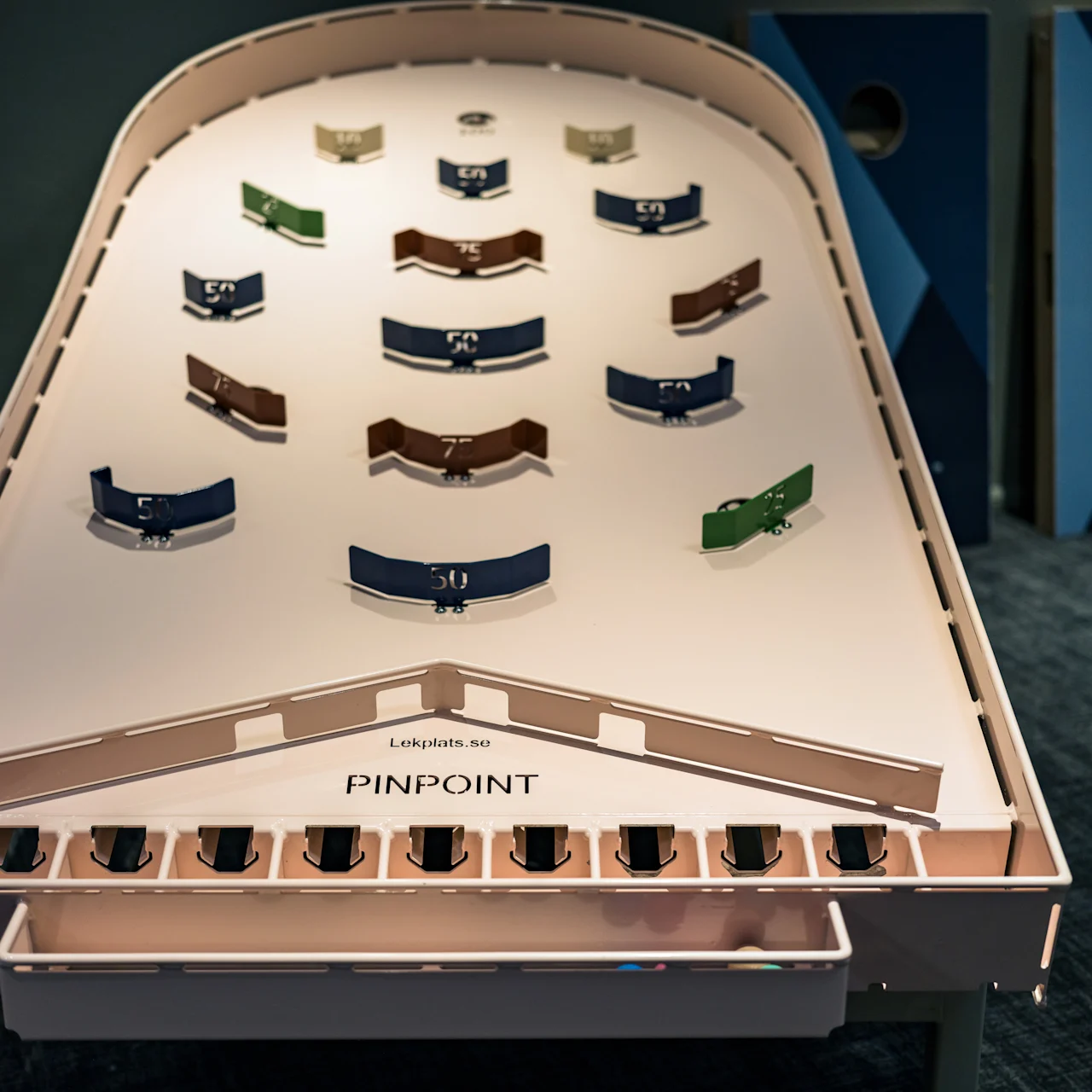 A pinball-style table, designed for a game with colored flippers labeled with point values, sits in a room with carpet. Text on the table reads "Lekplats.no PINPOINT."