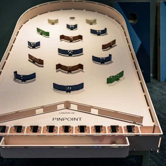 Et flipperspill-lignende bord, designet for et spill med fargede flippere merket med poengverdier, står i et rom med teppe. Tekst på bordet lyder "Lekplats.no PINPOINT."
