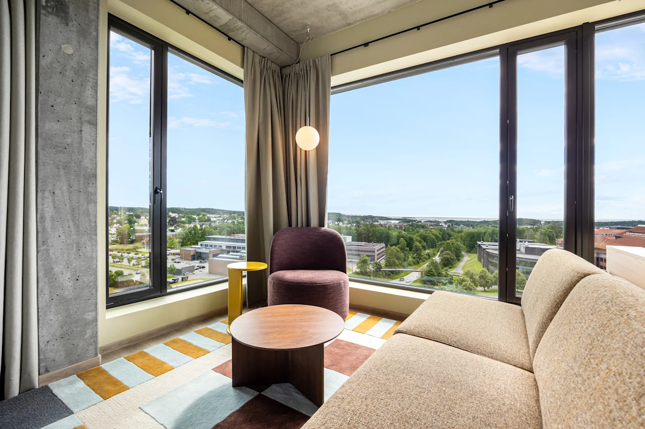 A modern hotel room with a comfortable sofa and armchair, featuring large windows that offer a panoramic view of the city.