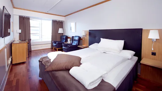 En pænt redt seng i et rent og moderne hotelværelse med træmøbler og naturligt lys fra et vindue.