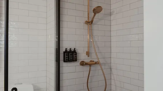 Et stilfuldt bruseområde med hvide fliser, elegante guldfarvede armaturer og praktiske vægmonterede dispensere.
