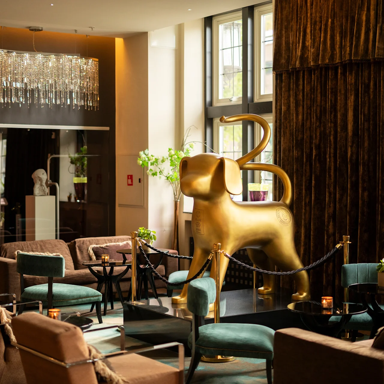 A large golden dog sculpture stands on a black platform, surrounded by seating and tables, in a luxurious room with chandelier and velvet curtains.