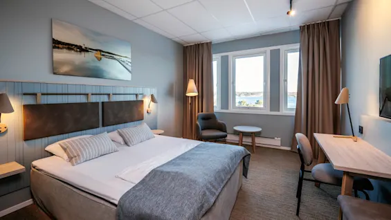 A neatly arranged hotel room features a large bed with grey accents, a desk, chair, and a window revealing a scenic view, adorned with modern lamps.