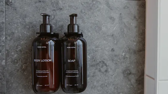 Kaksi tummaa pumppupulloa harmaalla kaakeliseinällä, merkitty BODY LOTION ja SOAP, henkilökohtaiseen hygieniaan.