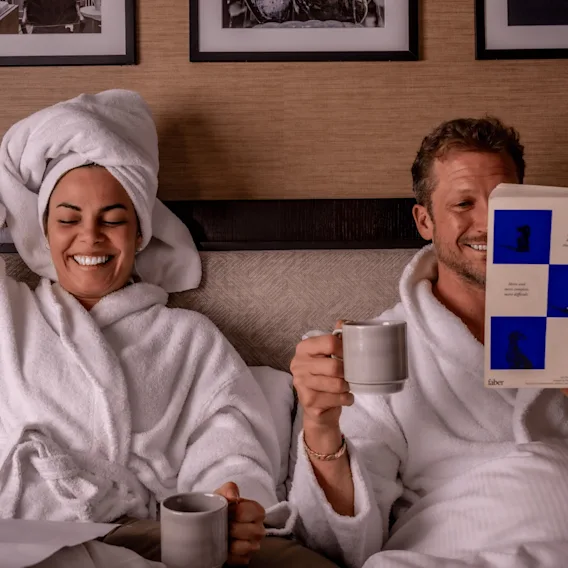 En man och en kvinna i vita badkappor kopplar av i en hotellsäng. Kvinnan har en handduk virad runt huvudet och skrattar, medan mannen håller i en mugg och läser boken "Intermezzo" av Sally Rooney.