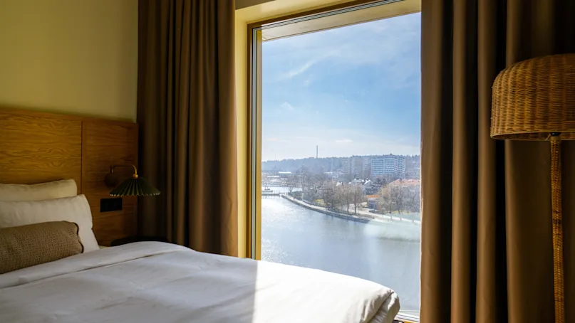 A neatly made bed is illuminated by sunlight streaming through a large window, overlooking a river and distant buildings on a clear day.