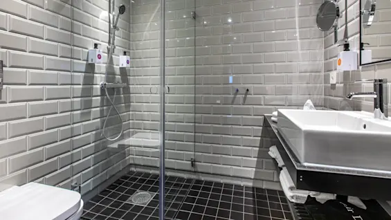 A glass shower enclosure with stainless steel fixtures and soap dispensers is in a modern bathroom. Gray subway tiles cover the walls, with a white sink on a black counter.