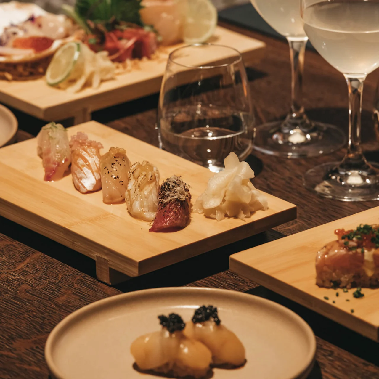 Assortert sushi på treplanker, elegant presentert ved siden av fylte vinglass på en mønstret bakgrunn.