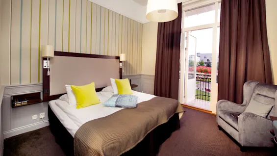 A bed with brown covers and yellow pillows sits against a striped wall, next to an open balcony door with curtains, overlooking a garden. An armchair is nearby.
