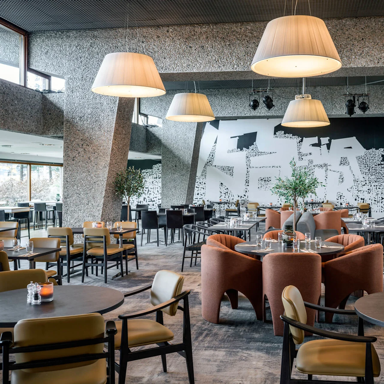 Modern dining area with round tables and chairs, under large hanging lamps. Artistic wall mural and scattered plants enhance the contemporary, open-plan restaurant setting.